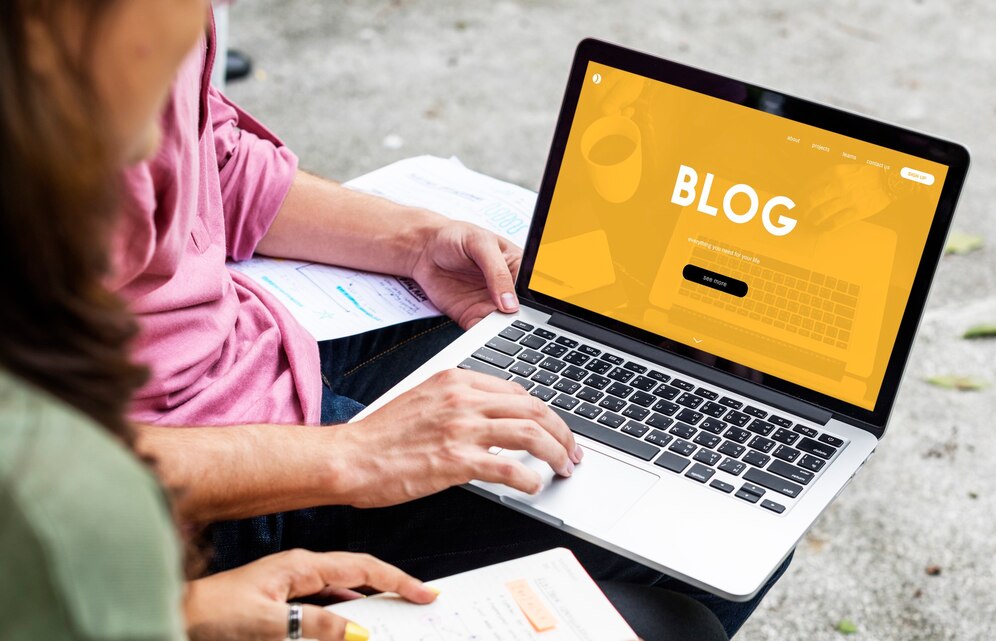 Two people collaborating, one working on a laptop with a bright yellow blog page displayed, featuring the word 'BLOG' in large white letters, possibly discussing guest posting strategies. The other person is holding a notebook and taking notes.